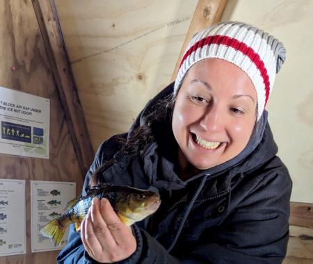 A woman is inside a wooded fish hut with a winter hat and coat. She smiles holding a fish.